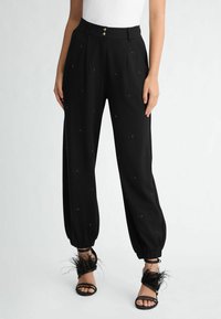 Pantaloni neri plissettati con dettagli testurizzati e polsini elastici, dotati di due bottoni frontali. Indossati con sandali alla schiava decorati con piume.