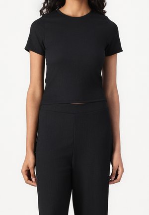 Femme portant un crop top noir moulant à manches courtes et un pantalon noir taille haute assorti, sur un fond uni.