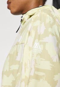 Ανοιχτό κίτρινο μπουφάν με υφή και αφηρημένο μοτίβο. Διαθέτει φερμουάρ μπροστά, κουκούλα με κορδόνι και λευκό λογότυπο στο μανίκι.
