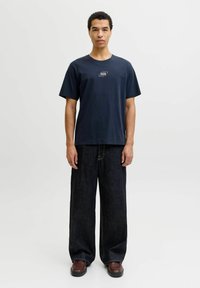 Camiseta azul marino con cuello redondo que presenta un pequeño parche con logo. Combinada con jeans de mezclilla oscura de pierna ancha y zapatos marrones. Diseño sencillo, con detalles mínimos.