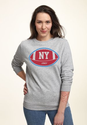 Femme portant des boucles d'oreilles, un sweat-shirt gris avec un motif de football rouge intitulé "NY 1961", un jean bleu, et montrant un tatouage floral sur son avant-bras gauche.