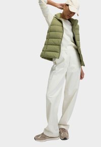 Olijfgroen puffer vest met een hoge kraag, crèmekleurig langemouwen shirt, witte wijde broek en bruine sneakers met beige accenten.