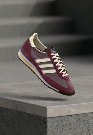 Baskets bordeaux et beige avec des rayures blanches et des lacets flottant au-dessus d'un rebord en béton sur un fond d'escaliers floutés.