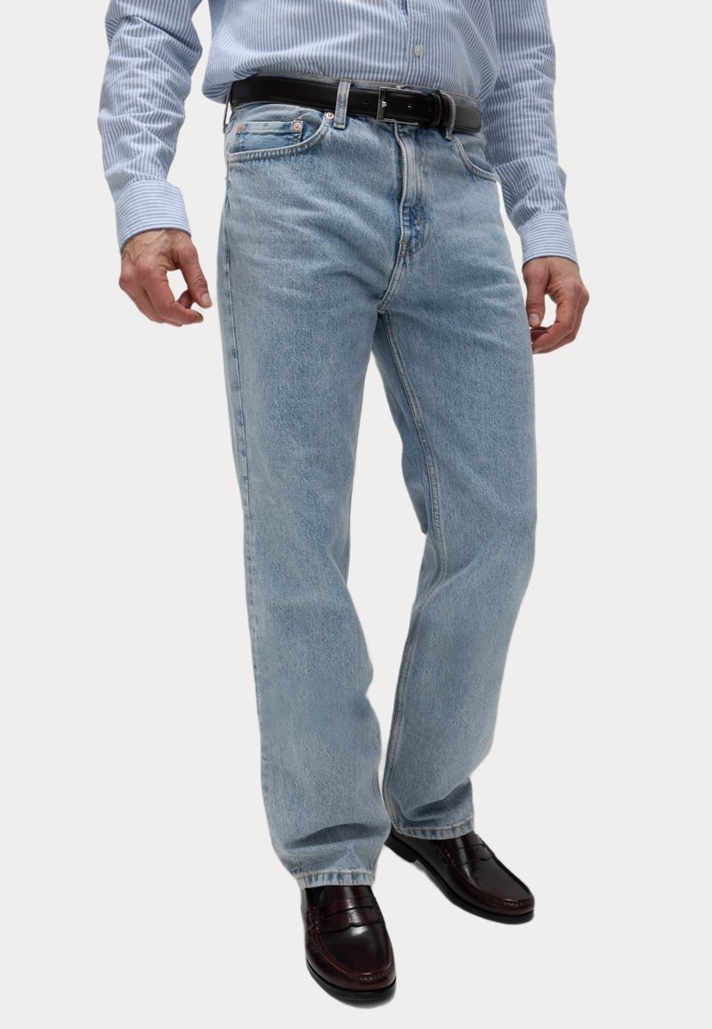 Vaqueros de denim azul claro de corte recto, con trabillas para cinturón y cremallera. Llevados con una camisa a rayas y mocasines oscuros.