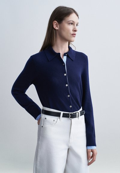 Femme aux longs cheveux bruns portant un cardigan bleu marine boutonné par-dessus une chemise bleue, un jean blanc taille haute et une ceinture noire, regardant vers la droite.