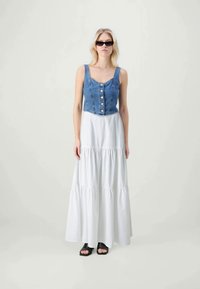 Top de corpete em denim com detalhes em botão, combinado com uma saia maxi branca e em camadas. Calçado aberto preto completa o conjunto.