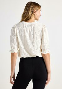 Kvinde set bagfra iført en hvid tekstureret bluse med ballonærmer og sorte tætsiddende bukser mod en ensfarvet baggrund.