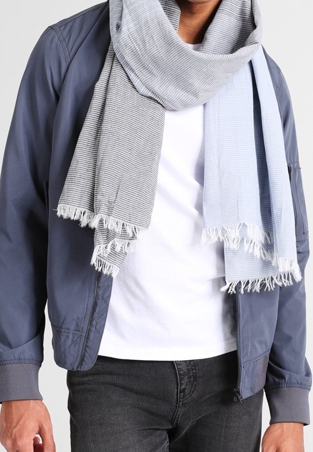 Homme portant une veste bleu marine, une chemise blanche, un jean noir et une écharpe rayée bicolore à franges drapée autour du cou.