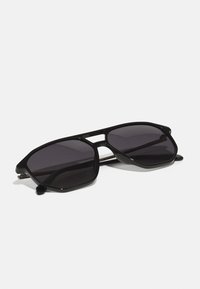 Lunettes de soleil noires avec une monture hexagonale, verres teintés foncés, design minimaliste et accents métalliques sur les branches.