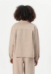 Giacca in corduroy beige con collo a revers, spalle cadenti e texture a costine. Presenta un giogo sul retro e polsini con bottoni per la chiusura.