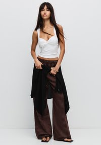 Hvid tætsiddende tanktop med snoet front, parret med brune stribede brede bukser og sorte slip-on sandaler. Sort skjorte bundet om taljen.
