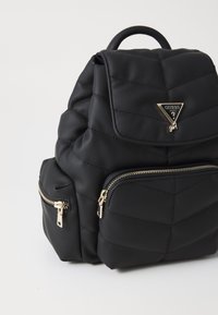 Sac à dos noir matelassé avec une plaque logo dorée, comprenant un compartiment principal et deux poches zippées à l'avant, fabriqué en matériau texturé.