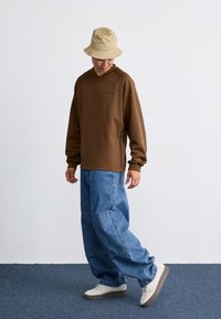 Brown long-sleeve sweatshirt with a v-neck, paired with loose blue denim jeans and beige bucket hat, worn with white sneakers.