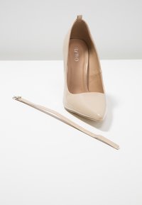 Beige lackläderhögklackad sko med spetsig tå, platt klack och ett fäst band. Slät textur med mjukt innertyg.