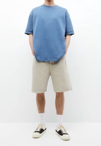 Hombre vistiendo pantalones cortos gris claro hasta la rodilla, camisa de manga corta azul y holgada, calcetines blancos y zapatillas negras con blancas, de pie con las manos en los bolsillos.