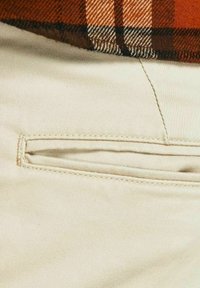 Beige katoenen broek met een klein, schuin zakje aan de zijkant met contrasterende stikdetails. De stof lijkt glad en licht textuur te hebben.