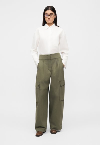 Chemise blanche à col et pantalon cargo vert olive taille haute avec poches latérales ; le mannequin porte des chaussures marron et des lunettes ; texture du tissu lisse.
