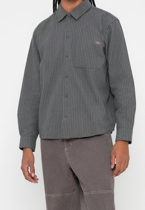 Homme portant une chemise grise à manches longues à boutons avec de fines rayures blanches verticales et une poche poitrine, associée à un pantalon marron atténué.