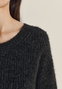 Tuvplāns cilvēkam, kurš valkā tumši pelēku, pūkainu adītu džemperi ar apaļu kakla izgriezumu uz gaiši bēša fona.