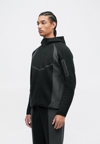 Czarna polarowa kurtka z kapturem zapinana na zamek, z ciemnoszarymi bocznymi wstawkami, wyposażona w kieszeń na suwak na rękawie oraz teksturowane wykończenie. Dopasowany krój z regulowanym dołem.