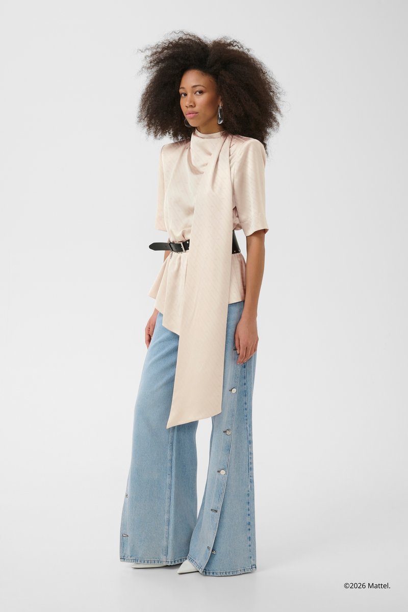 Kvinde med fyldigt krøllet hår iført en beige satinbluse med et langt bånd, sort bælte og lyseblå viddejeans med knapper i siden.