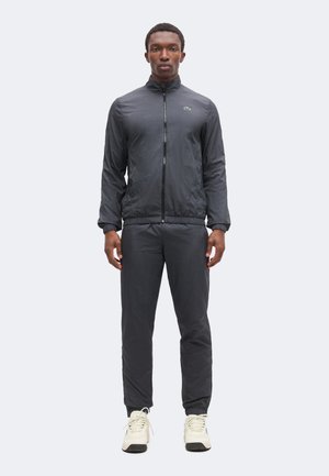 Hombre de pie mirando hacia adelante, vestido con una chaqueta de chándal negra con cremallera y pantalones, con zapatillas blancas, contra un fondo claro y liso.