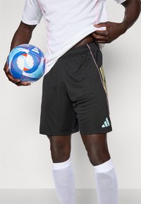 Svart treningsshorts med snøring i livet, med grønne striper og Adidas-logo. En blå og oransje fotball holdes i nærheten.