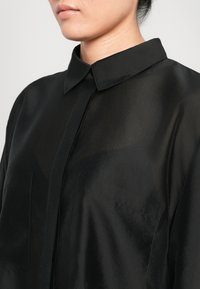 Nærbilde av en person som har på seg en semi-gjennomsiktig svart bluse med spiss krage og skjult knappestolpe mot en ensfarget bakgrunn.