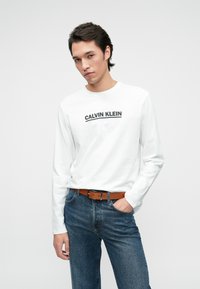 Bela srajca z dolgimi rokavi s črnim logotipom "Calvin Klein", kombinirana z modrimi traper hlačami in rjavim pasom. Preprost dizajn, sproščen kroj.