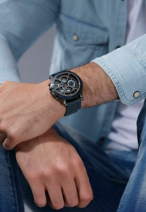 Man wearing blue denim shirt and jeans shows black and blue analog wristwatch with textured rubber strap on left wrist.
