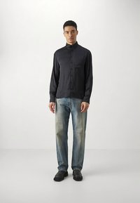 Jonge man staand, gekleed in een zwart overhemd met knopen, vervaagde blauwe jeans met kniekappen en zwarte schoenen tegen een effen witte achtergrond.