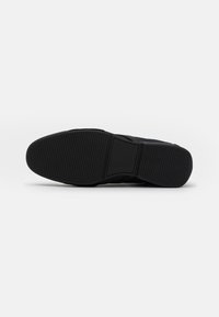Schwarze Sneakersohle aus strukturiertem Gummi, mit einem geriffelten Muster und Logo. Das Design ist flach mit einer abgerundeten Spitze und minimalistischem Stil.