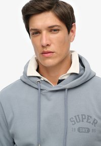 Sudadera de color azul claro hecha de algodón suave. Cuenta con un capucha con cordones, un cuello interior color crema y un detalle impreso en el pecho que dice "SUPER DRY 1985".