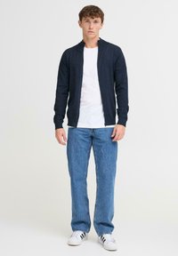 Tengerészkék kötött kardigán cipzárral, fehér póló fölött viselve. Világoskék egyenes szárú farmerrel és fehér sportcipővel párosítva.