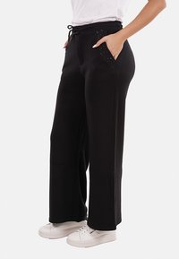Donna che indossa pantaloni neri a gamba larga con tasche frontali decorate, sneakers bianche e una maglietta bianca, in piedi con le mani nelle tasche.