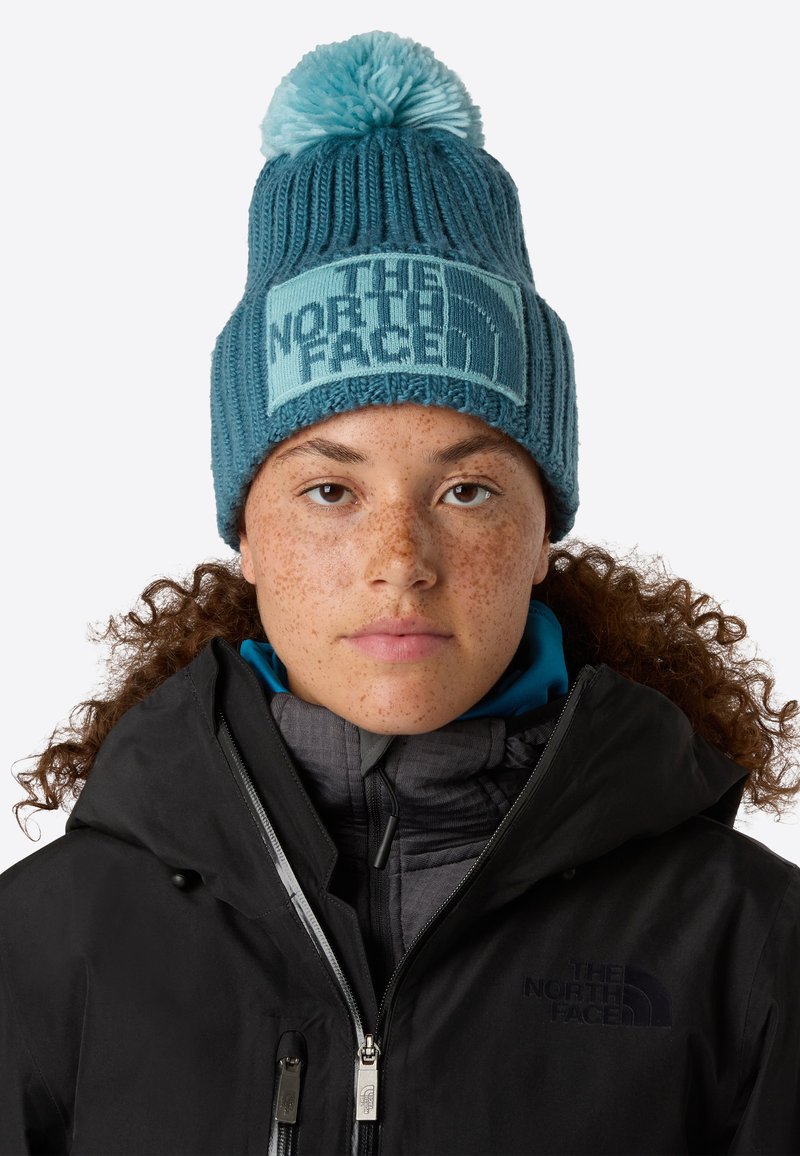 Beanie lavorato a maglia in teal con una fascia con logo blu chiaro e pom-pom sopra. Texture a coste e vestibilità aderente, indossato con una giacca nera.