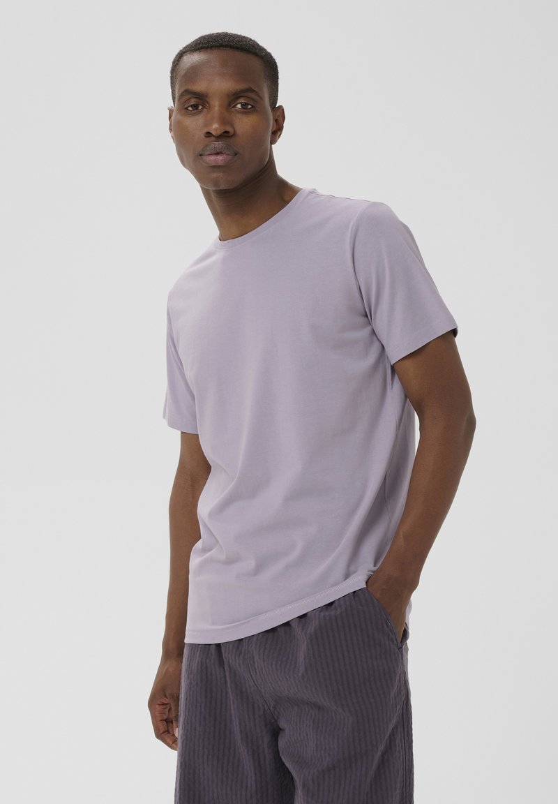 Homme portant une chemise à manches courtes violet clair et un pantalon sombre texturé, debout avec une main dans la poche, sur un fond uni.