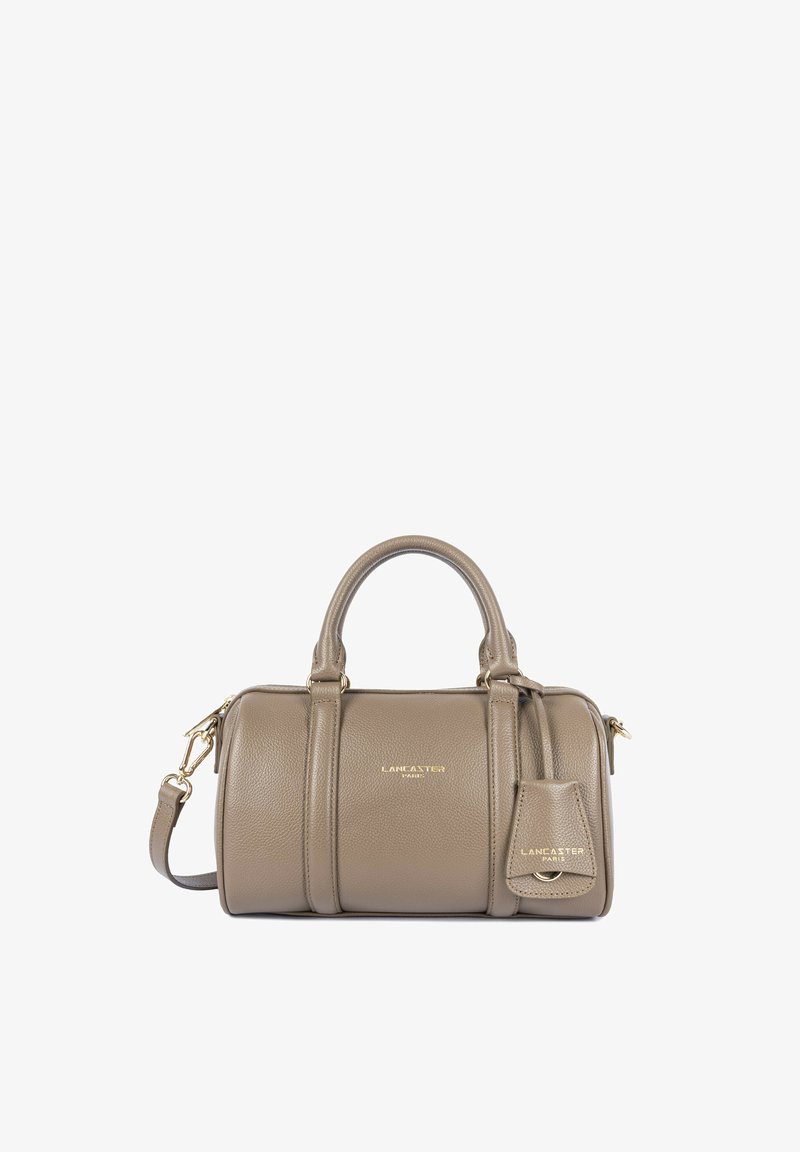 Sac seau en cuir taupe avec deux anses, une bandoulière détachable et une petite étiquette à bagages assortie, griffé Lancaster Paris en texte doré.