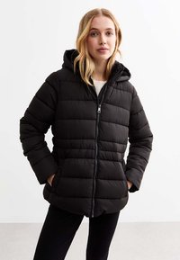 Doudoune noire avec capuche, présentant une texture matelassée, une fermeture éclair à l'avant et deux poches latérales. Portée par-dessus un haut de couleur claire.