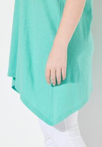 Tunic verde menta con una texture morbida, caratterizzata da un'orlo asimmetrico. Mano appoggiata sul fianco, abbinata a pantaloni bianchi.