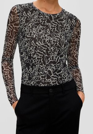 Zwart doorschijnend langarmshirt met wit abstract bloemenpatroon, gedragen met zwarte hoge taillebroek, handen in de zakken.