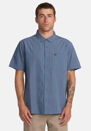 Mężczyzna o krótkich włosach, ubrany w niebieską, krótką koszulę na guziki z cienkimi czerwonymi pionowymi paskami oraz beżowe spodnie, stoi na białym tle.