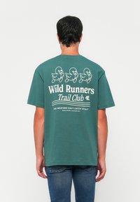 Muškarac okrenut leđima nosi zelenu majicu s tri ilustracije trkaćih pataka i tekstom "Wild Runners Trail Club" i "Vrijeme nas ne može uhvatiti".
