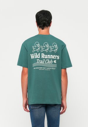 Mann, der sich abwendet, trägt ein grünes T-Shirt mit drei laufenden Entenillustrationen und dem Text "Wild Runners Trail Club" sowie "Das Wetter kann uns nicht einholen."