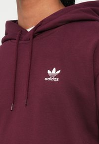 Közelkép egy személyről, aki bordó Adidas kapucnis felsőt visel, amelyen a fehér Adidas logó és a zsinórok láthatóak a mellkas területén.