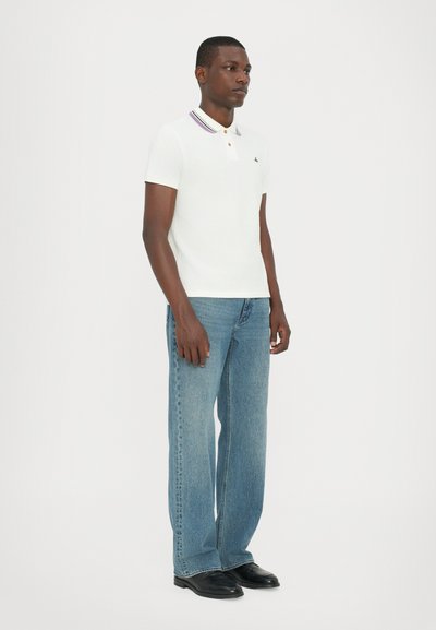 Balta polo krekla ar purpura svītraina apkakli, oranžu pogu, savienots ar gaiši zilu platu kāju biksēm un melniem apaviem. Modelis stāv sānis.