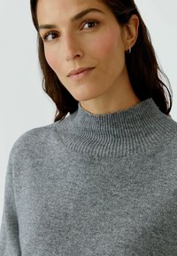 Pull en maille grise avec un col haut à côtes et une texture subtile. Le tissu semble doux et chaleureux, avec une coupe décontractée.