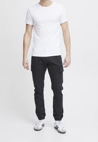 Uomo che indossa una semplice t-shirt bianca a maniche corte, pantaloni cargo neri con tasche laterali e sneakers bianche con strisce nere.