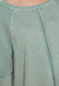 Világos türkiz oversized ing, egy texturált panellel az egyik oldalon és sima felülettel, kerek nyakkivágással és látható varrási részletekkel.