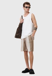 Weißes geripptes Tanktop, beige gestreifte Shorts, schwarze Slipper und eine dunkle gemusterte Tasche; accessoriert mit grauen Sonnenbrillen.